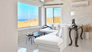 A modern living room with a white sofa, a small round table, and a view of the desert through large windows. - Luxurious villa with a swimming pool, sea and sunset view (Kéa)