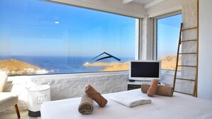 A bedroom with a large window overlooking a coastal view, a bed with rolled towels, a television, and a ladder against the wall. - Luxurious villa with a swimming pool, sea and sunset view (Kéa)