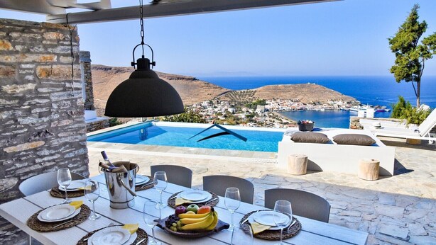 A dining table set with plates, glasses, and a fruit bowl, overlooking a pool and a coastal town. - Luxurious villa with a swimming pool, sea and sunset view (Kéa)