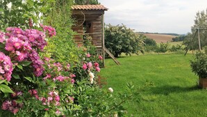 A rustic wooden cabin with a sloped roof, surrounded by lush greenery and flowering plants.