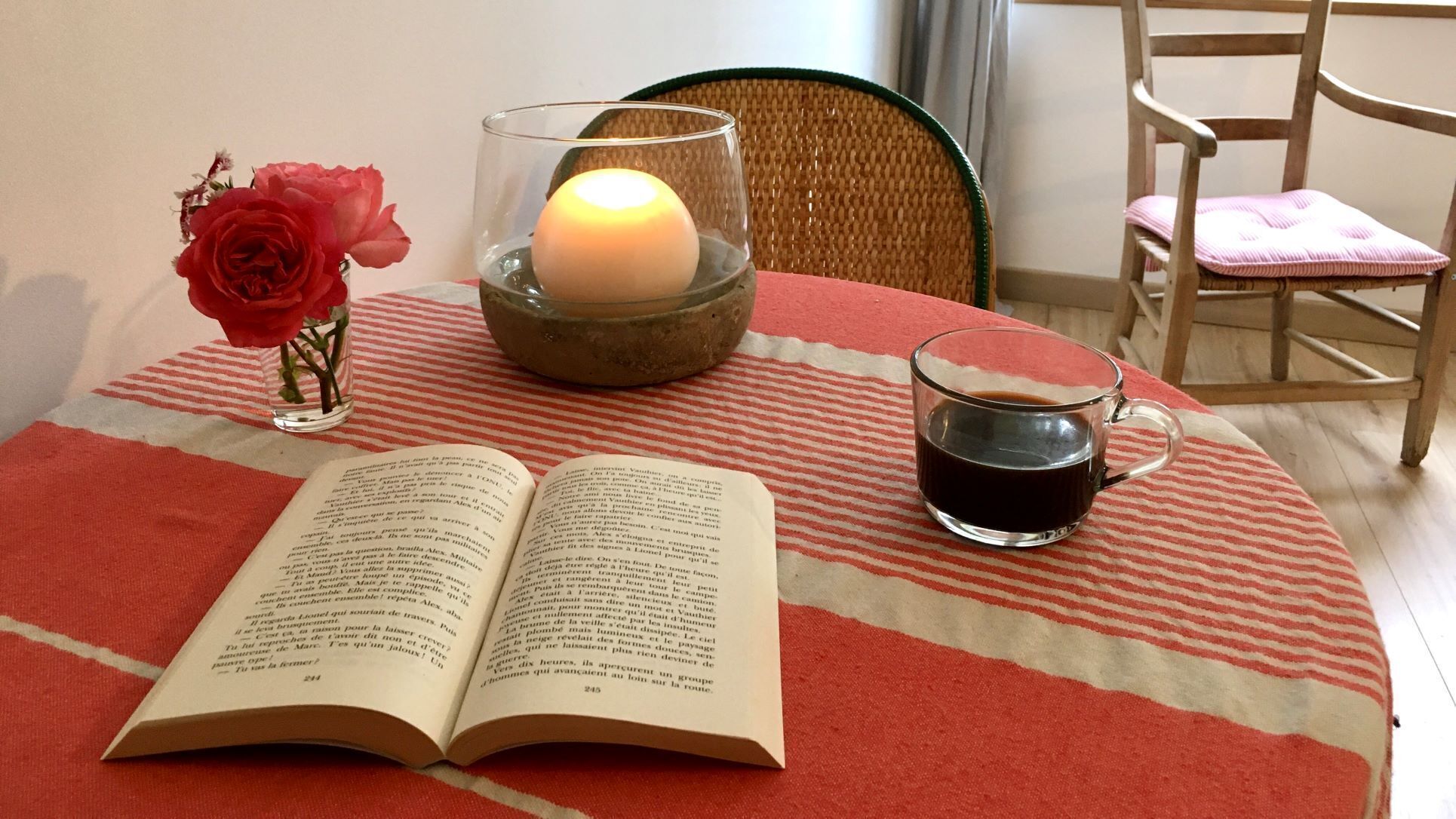 An open book, a candle, and a cup of coffee on a striped tablecloth.