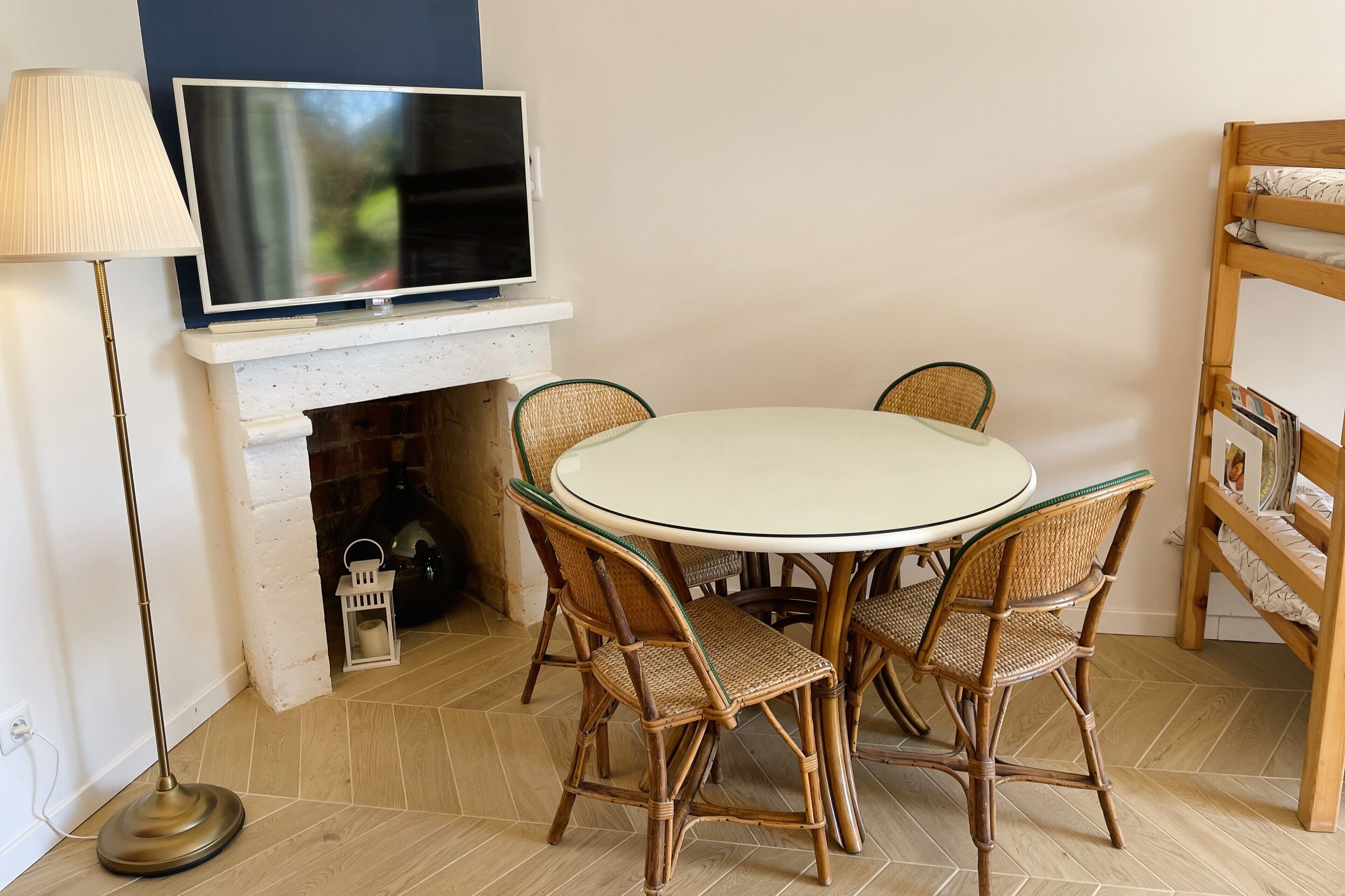 A dining area with a round table, wicker chairs, a flat-screen TV above a white mantelpiece, and a bookshelf to the right.