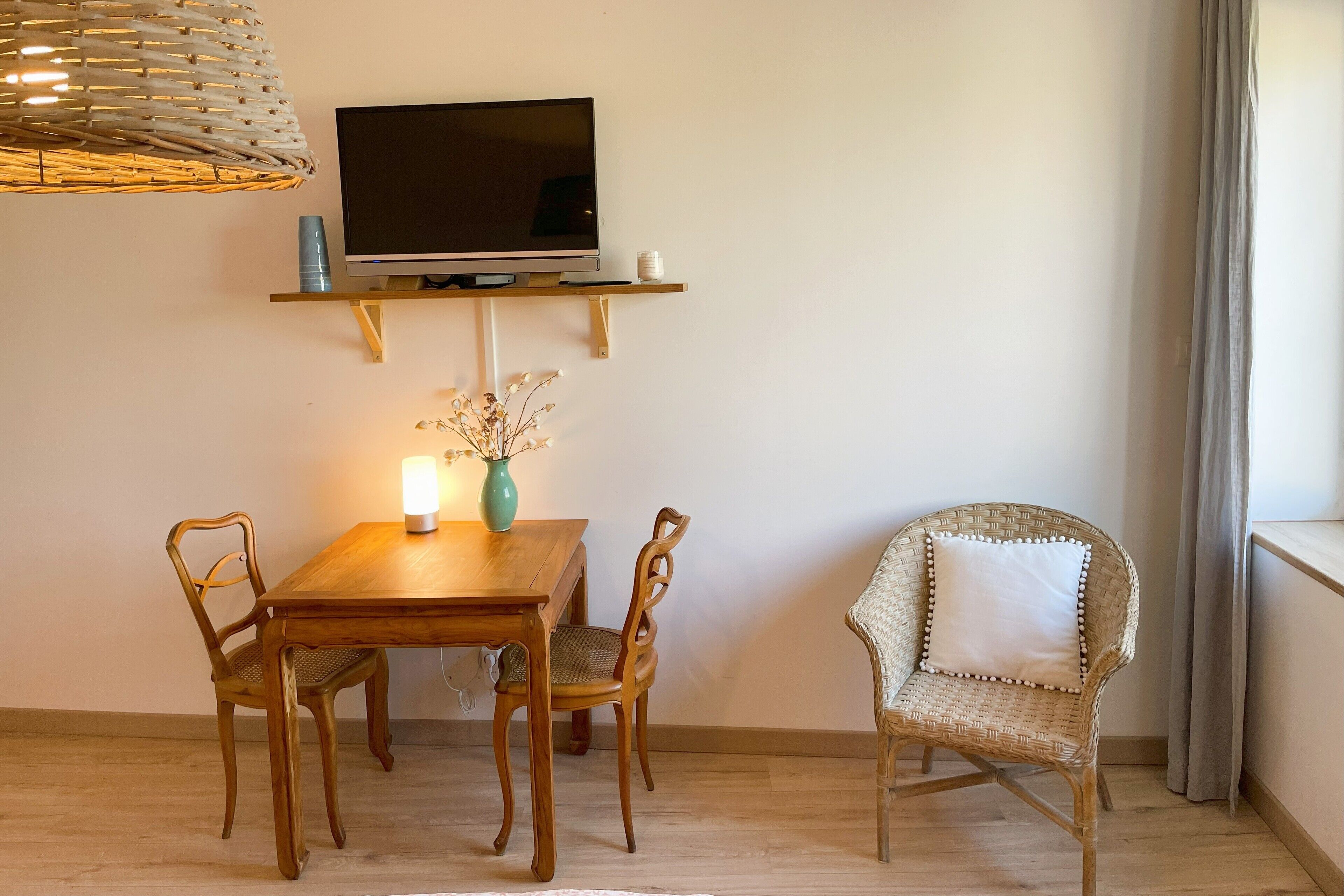 A dining area with a wooden table, two chairs, a TV on a shelf, a vase with dried flowers, and a wicker chair.
