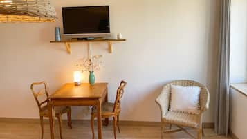 A dining area with a wooden table, two chairs, a TV on a shelf, a vase with dried flowers, and a wicker chair.