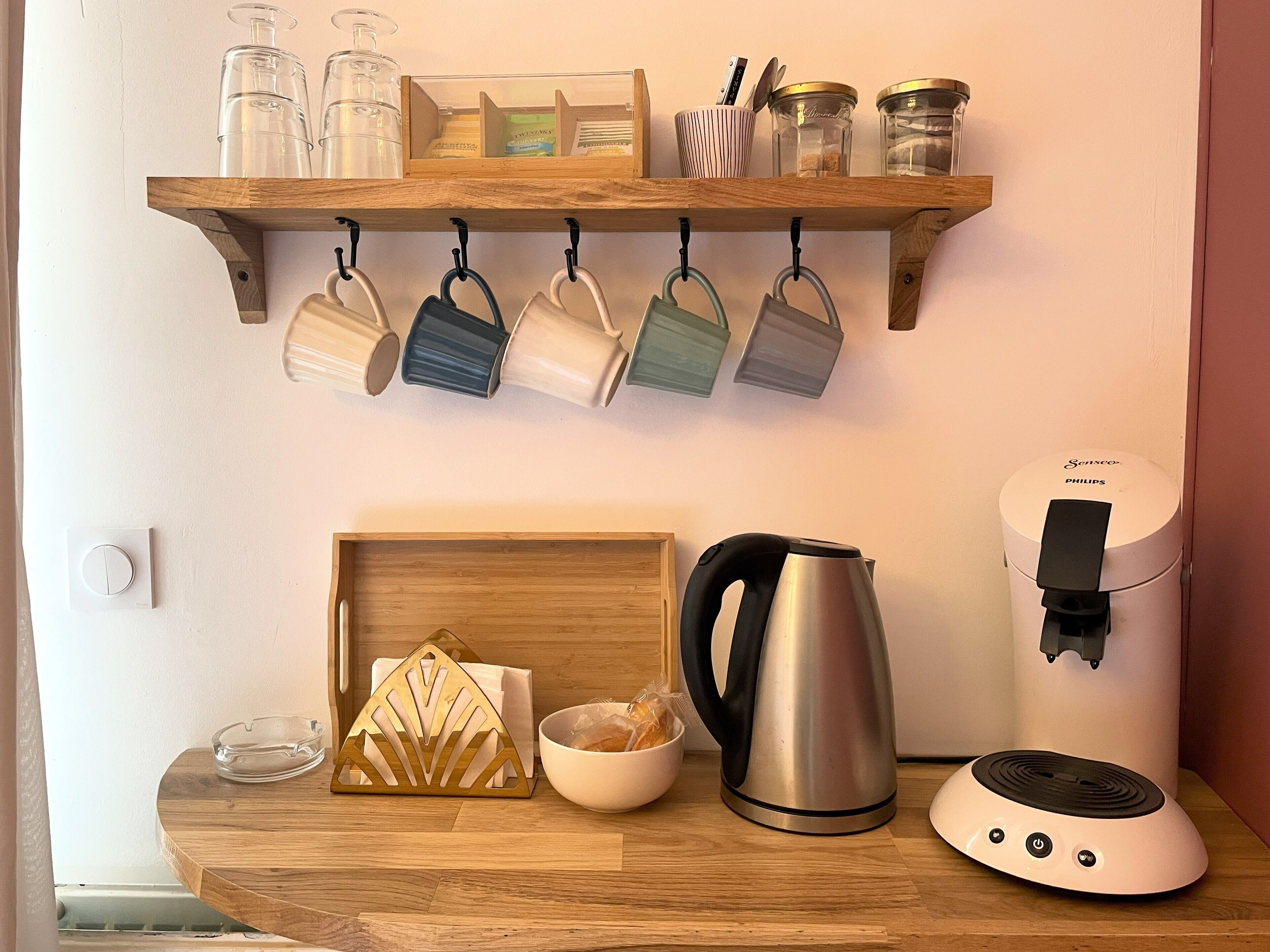 A kitchen counter with a kettle, a coffee maker, and a shelf above holding mugs and jars.