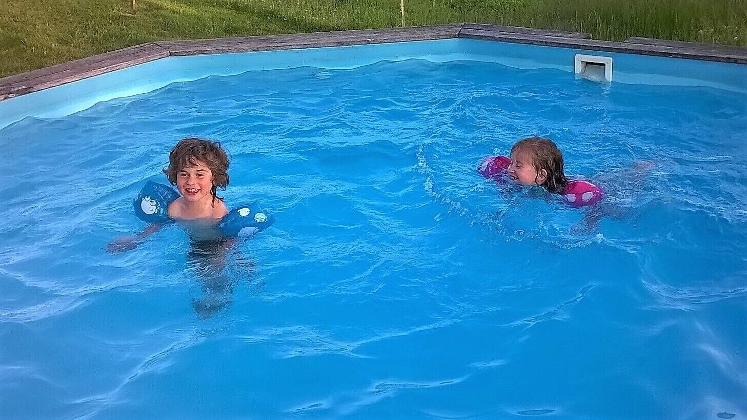 Two children in a swimming pool with a wooden deck and grass in the background.