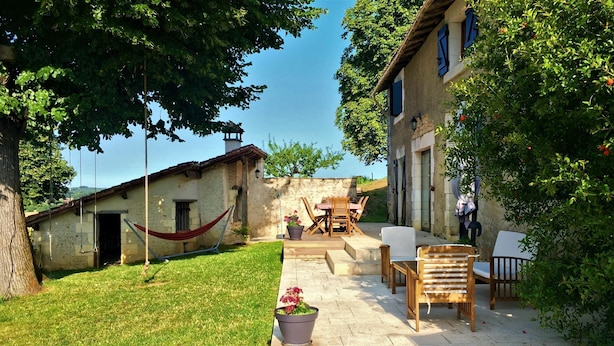 A rustic outdoor patio with a hammock, wooden furniture, and a stone building.
