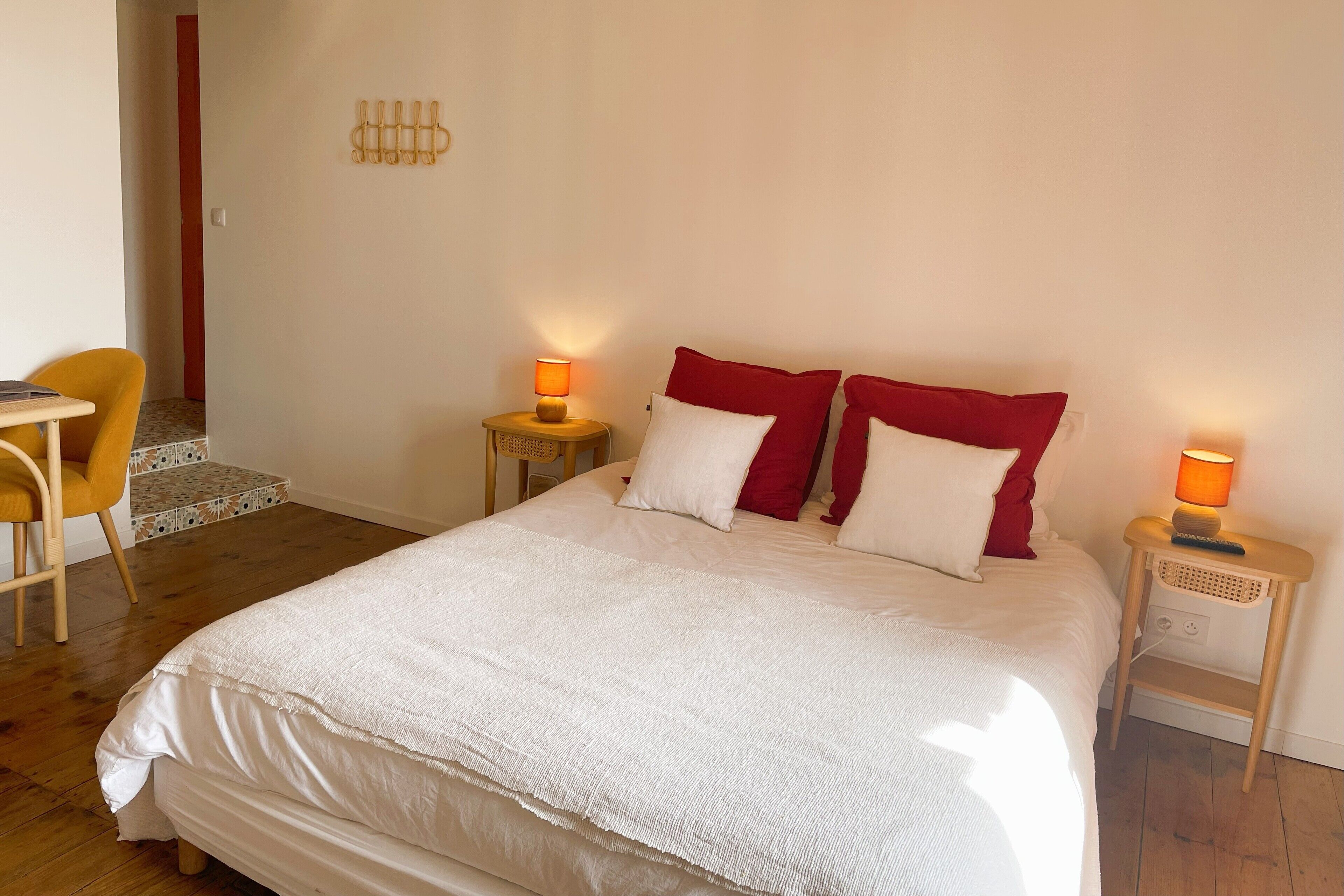 A neatly made bed with white and red pillows, a wooden nightstand with a lamp, and a yellow chair.