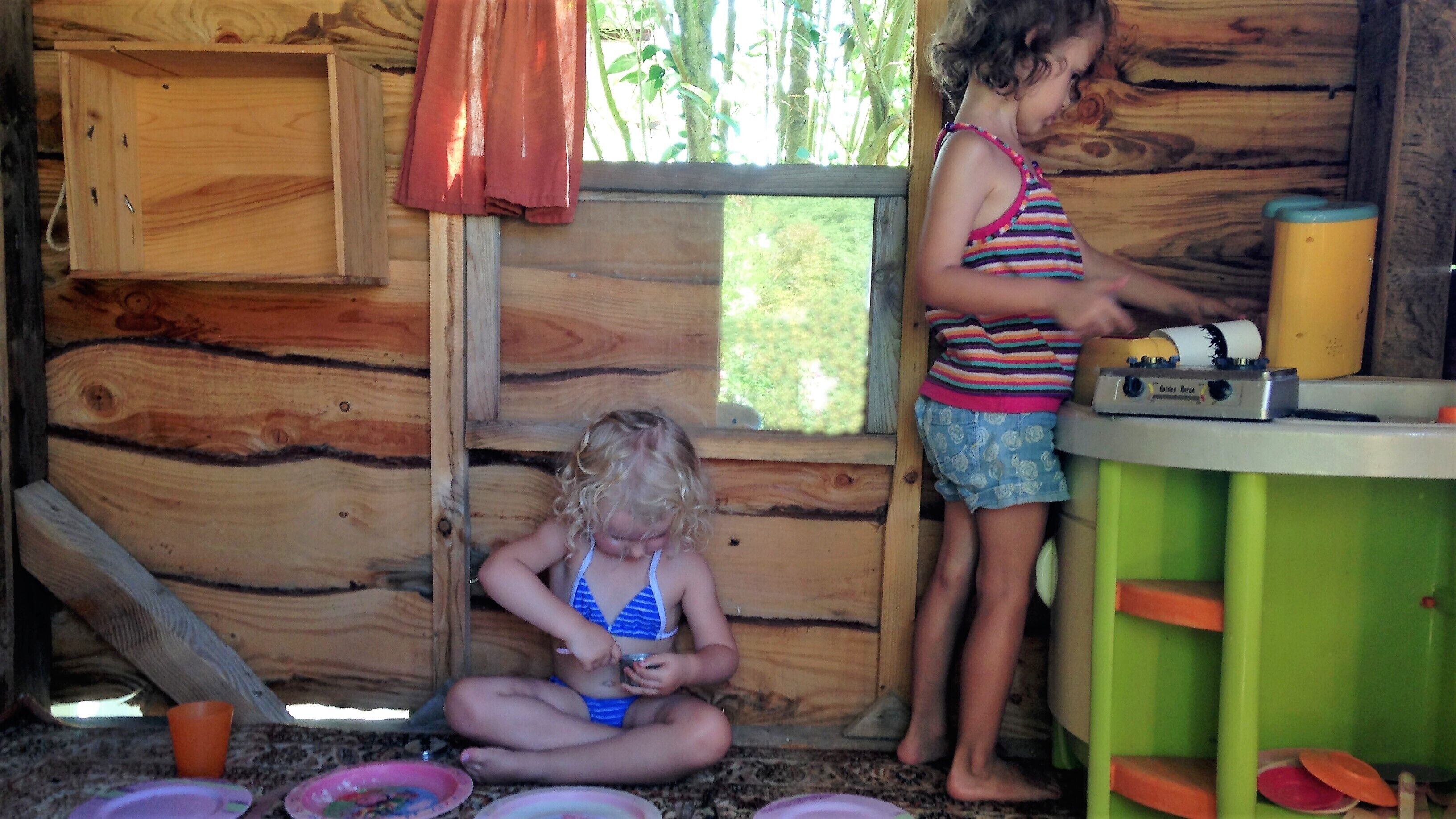Interior rústico de una cabaña de madera con dos niños jugando. Un niño está sentado en el suelo, mientras que el otro está de pie cerca de una mesa con una radio.