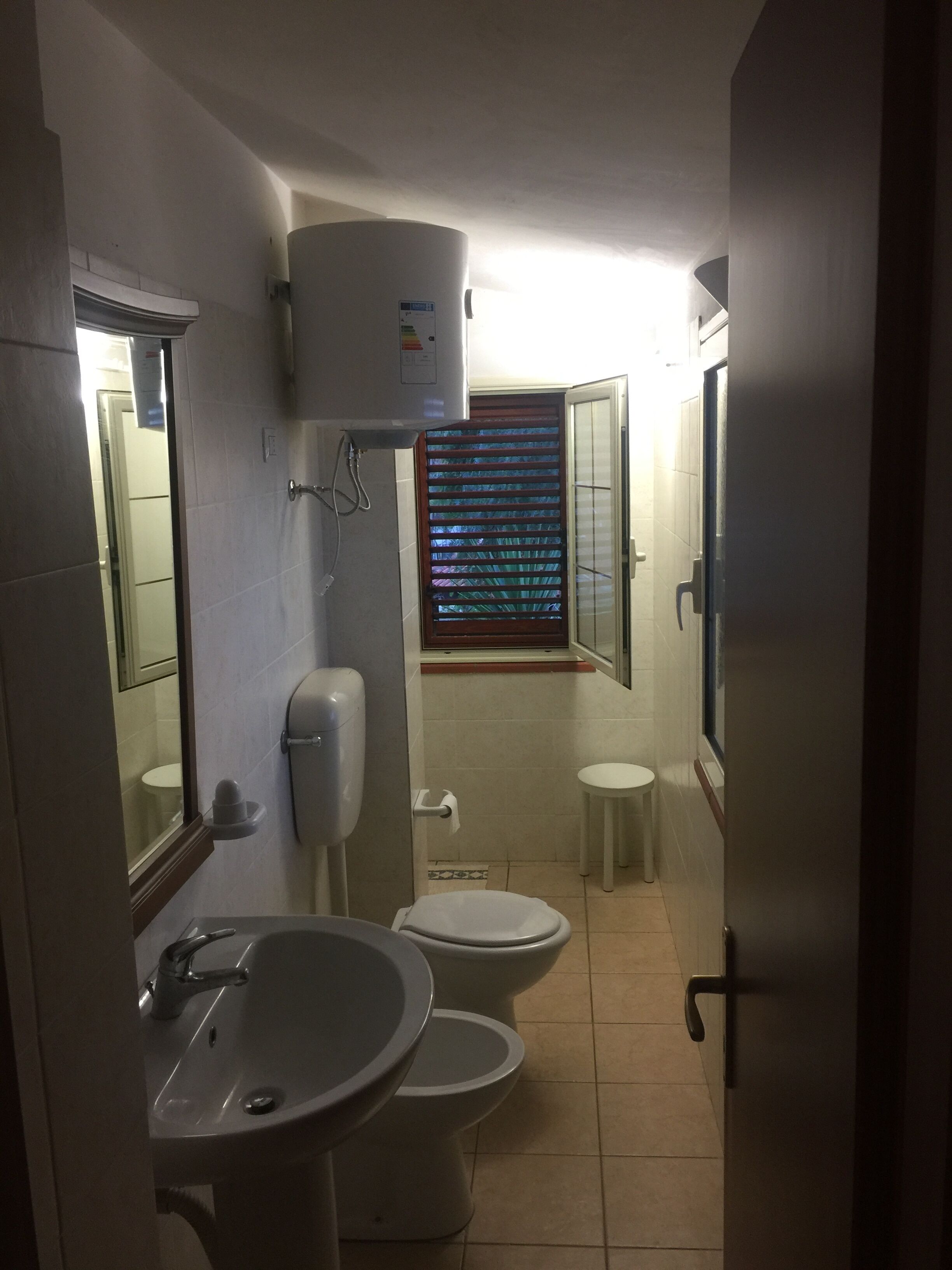 A compact bathroom with a sink, two toilets, a window with blinds, and a ceiling-mounted air conditioner.