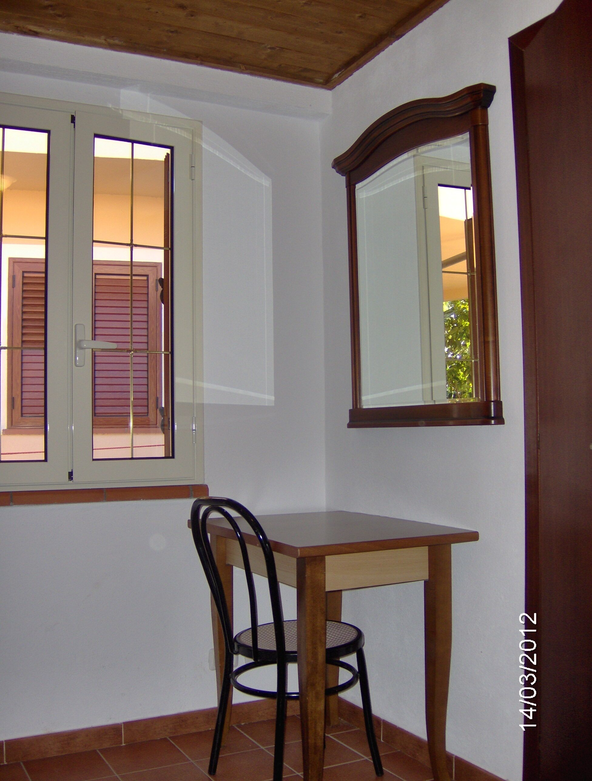 Une pièce avec une table en bois et une chaise, une fenêtre avec des stores et un miroir au mur.