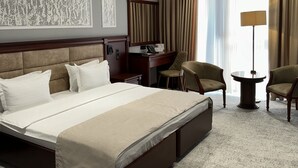 A hotel room with a large bed, a desk, two chairs, a lamp, and a window with curtains.