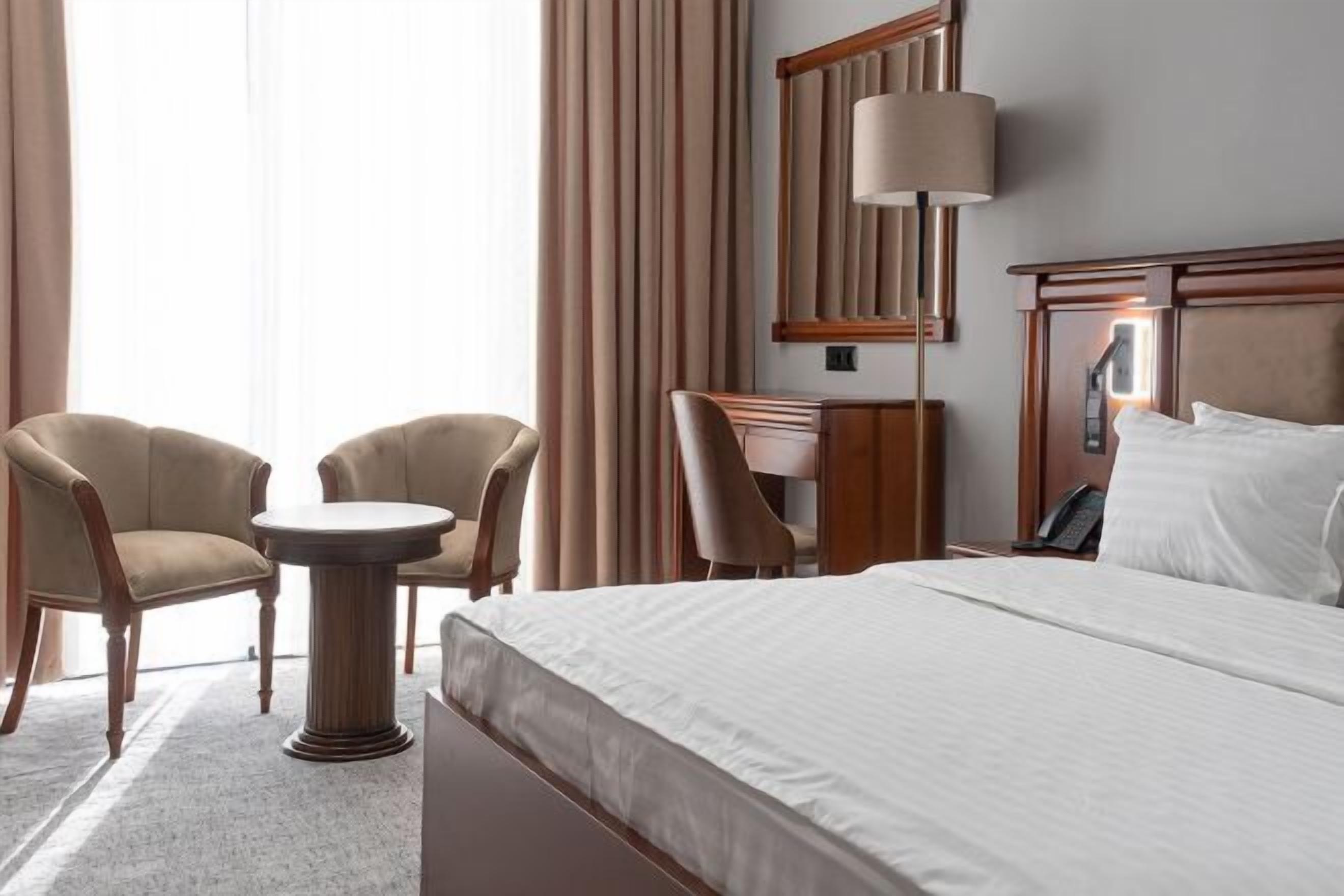 Ein Hotelzimmer mit einem Bett, zwei Sesseln, einem kleinen runden Tisch, einem Schreibtisch, einer Lampe und einem Spiegel.
