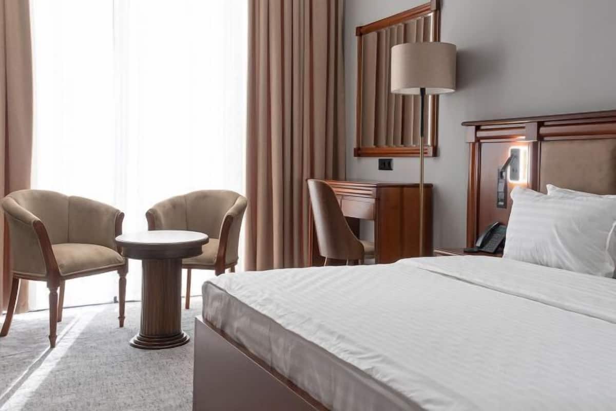 Ein Hotelzimmer mit einem Bett, zwei Sesseln, einem kleinen runden Tisch, einem Schreibtisch, einer Lampe und einem Spiegel.