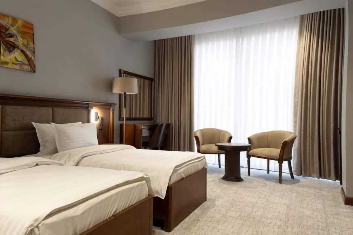Ein Hotelzimmer mit zwei Betten, einem Schreibtisch, einem Stuhl, einer Lampe und einem großen Fenster mit Vorhängen.