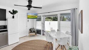 A modern kitchen with white cabinets, a built-in oven, and a microwave. There is a dining area with a white table and chairs, a large window, and a ceiling fan. - Spa,Fire Pit,Cinema - The Cottage Coolum Beach (Coolum Beach)