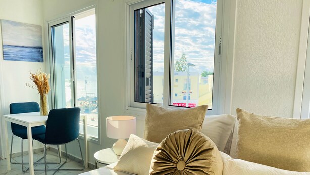 A hotel room with a bed, a desk, and a chair. There is a window with a view of buildings and a beach. - Phaedrus Living Luxury Flat Lighthouse75 (Paphos)
