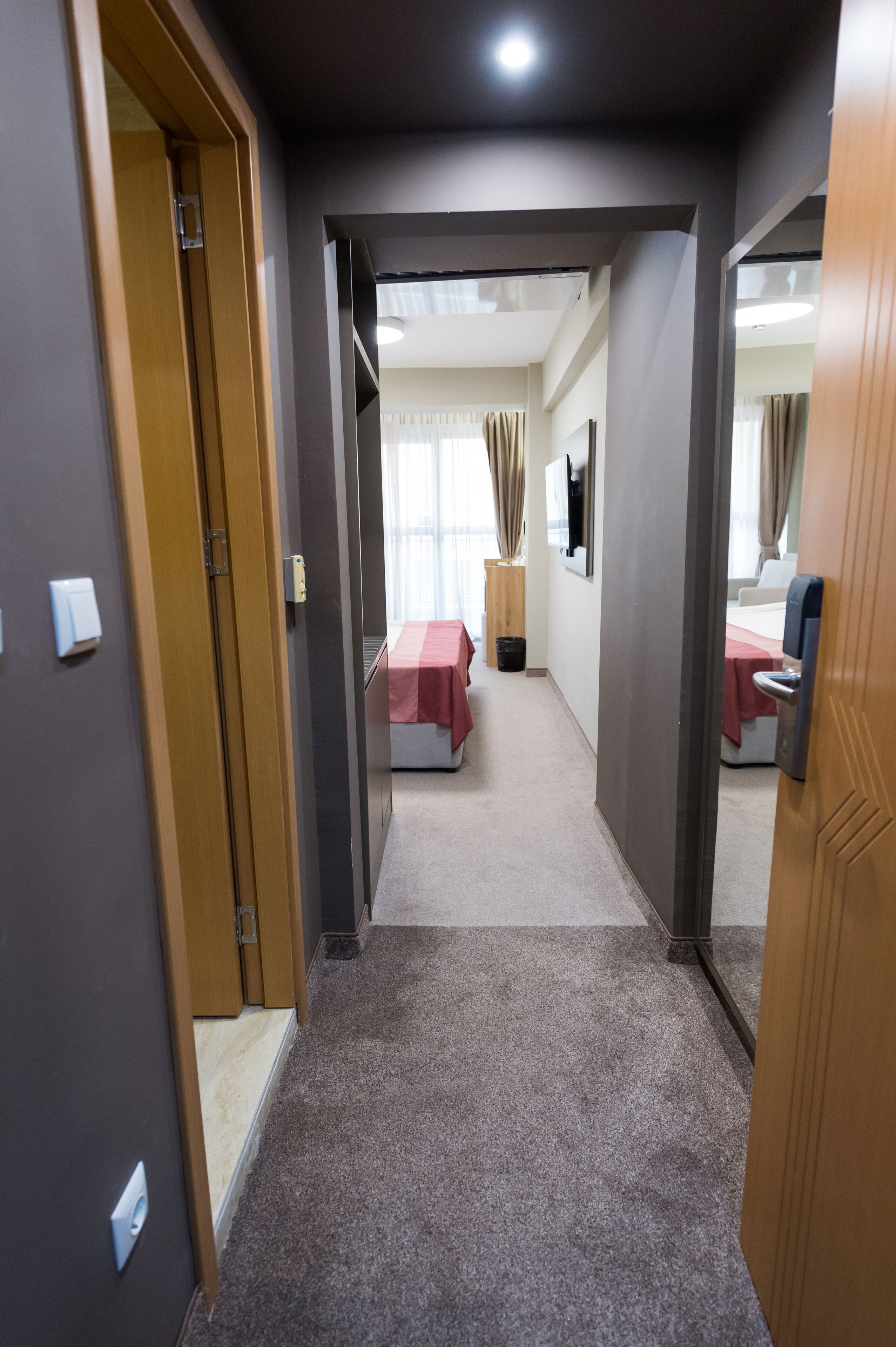 A hotel corridor with a carpeted floor, wooden doors, and a bed with a red bedspread in the background.