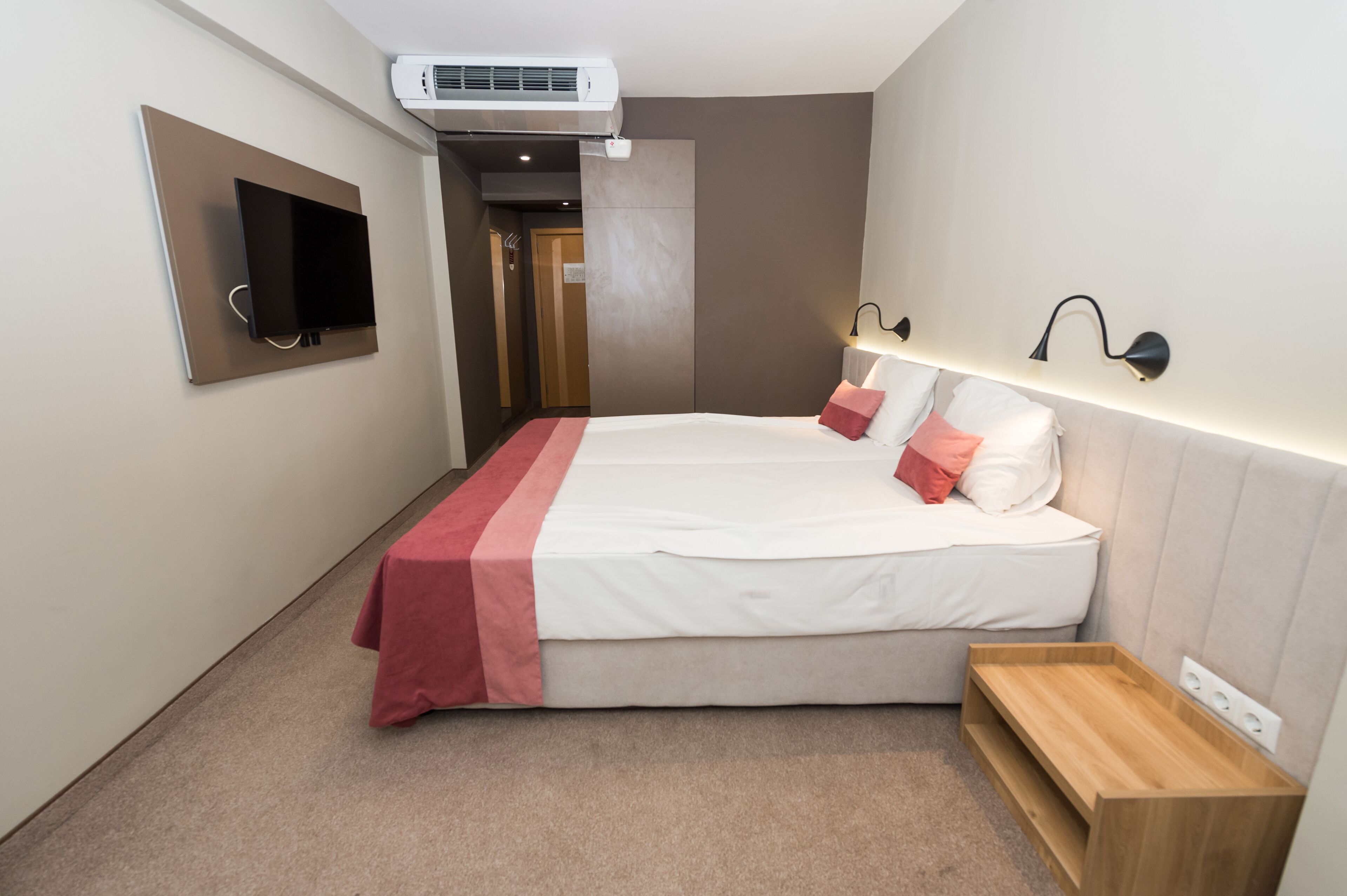 A hotel room with a bed, a TV mounted on the wall, a bedside table, and a wall-mounted lamp.