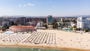 A beach with numerous sunbeds and umbrellas, a large building with a red roof, and a clear view of the ocean.