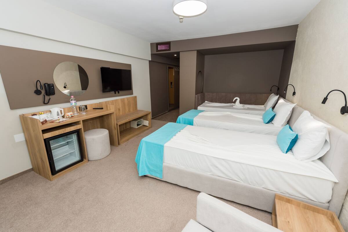 Habitación de hotel con dos camas individuales, un escritorio con computadora, un refrigerador pequeño, un espejo y un televisor montado en la pared.