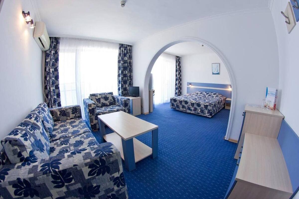 Habitación de hotel con sofá, una mesita, cama con colcha estampada de flores azules y blancas, y un ventanal con cortinas.