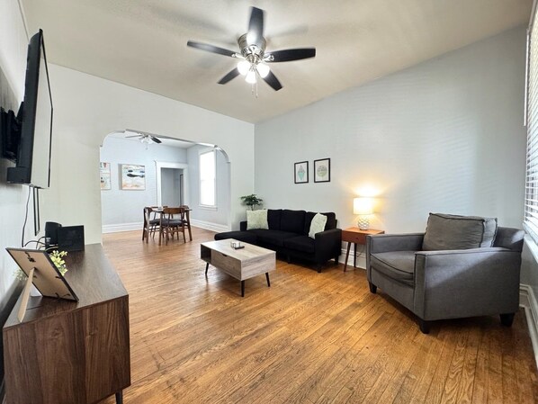 Una sala de estar moderna con ventilador de techo, un sofá negro, un sillón gris, una mesa de centro de madera y un televisor sobre un mueble bajo.