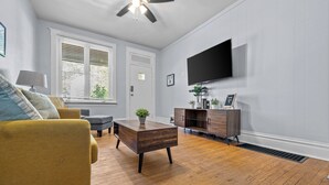 A living room with a yellow sofa, a wooden coffee table, a TV mounted on the wall, and a ceiling fan. - Dog-Friendly | Yard & Office (St. Louis)