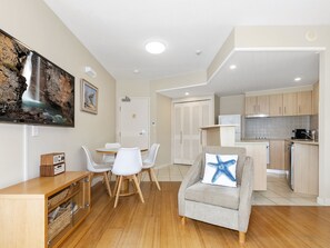A modern living room with a dining area, a kitchen, and a large TV mounted on the wall displaying a waterfall scene. - The Pool Resort Kingscliff - 2B Dual Key (6116/17) - Holiday Management Co. (Tweed)