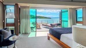 A modern hotel room with a large bed, a seating area with a view of the sea, and a balcony with a table and chairs.