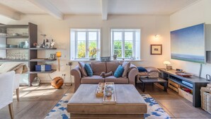Un salon confortable avec un canapé, une table basse et un téléviseur. Il y a deux fenêtres qui donnent sur un paysage verdoyant.