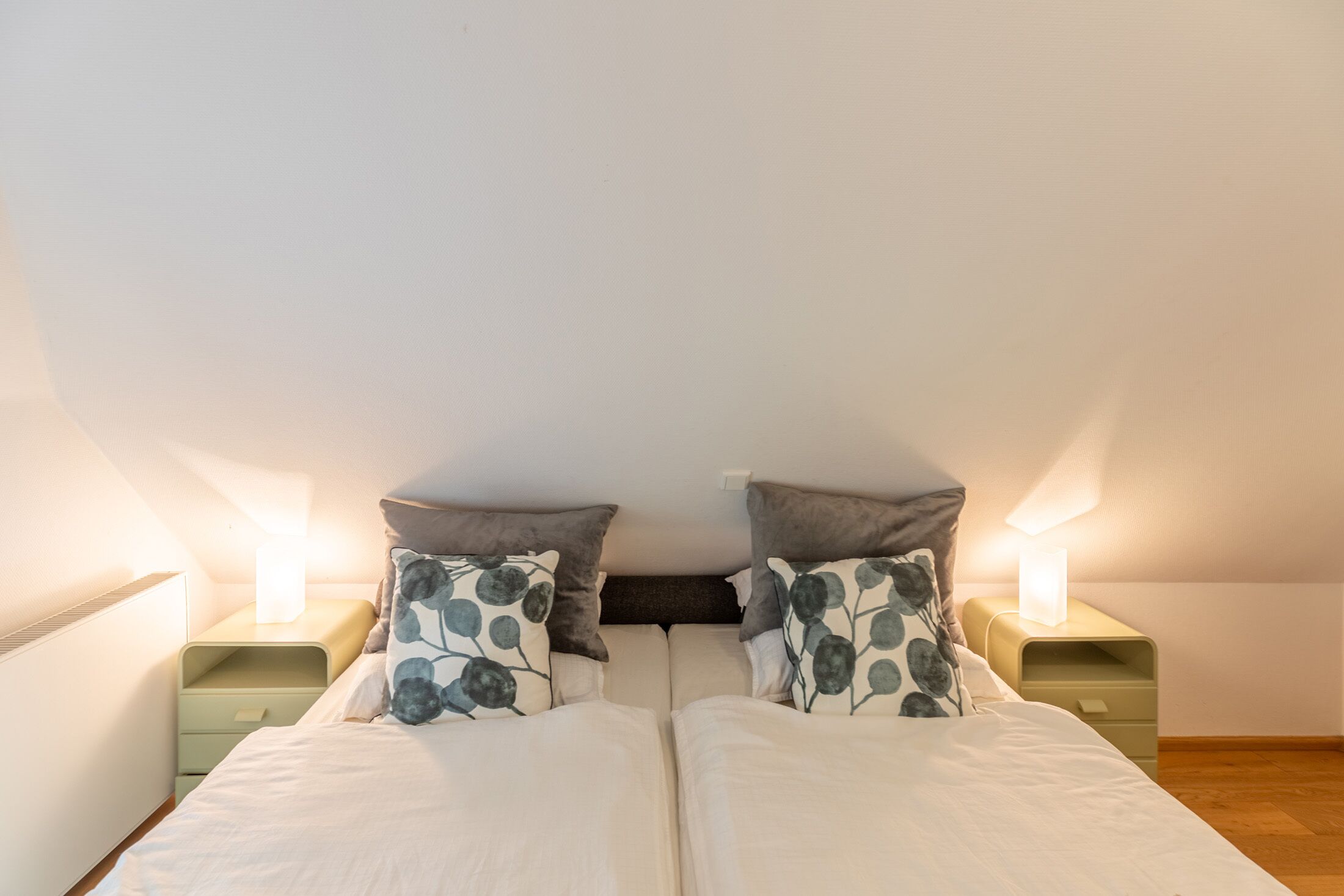 A twin bed with floral pillows, bedside tables with lamps, and a wooden floor.