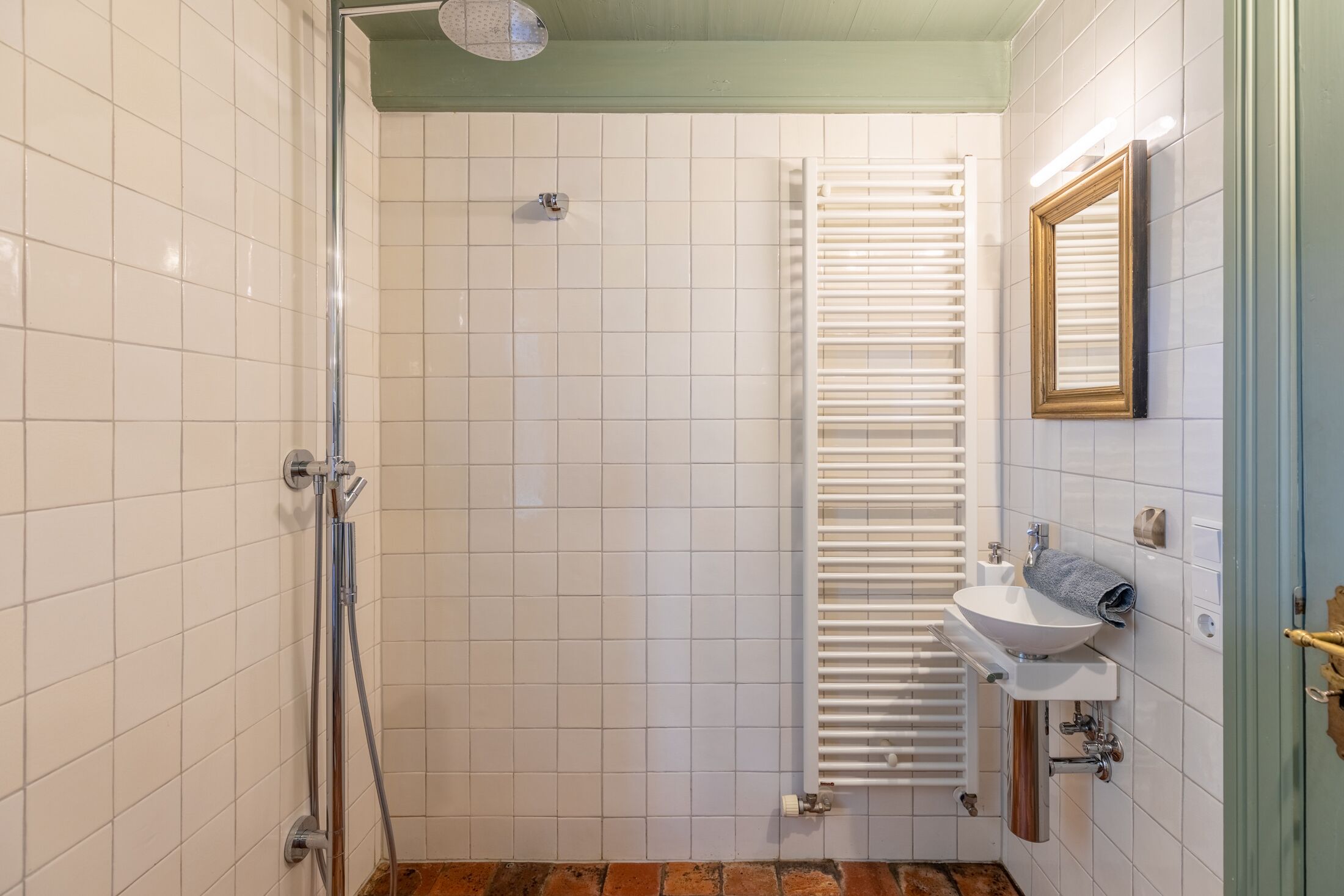 Un baño con paredes de azulejos blancos, una ducha con puerta de vidrio, un lavamanos blanco con una toalla y un espejo en la pared.