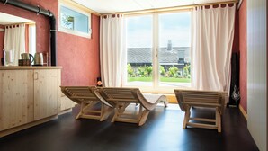 Una habitación con un banco de madera, una ventana con cortinas, vistas al exterior y una zona de cocina con armarios y electrodomésticos.