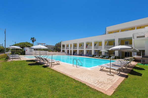 A swimming pool with sun loungers and umbrellas, surrounded by a well-maintained lawn and a building with multiple balconies.