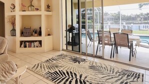Amplia sala de estar con un gran ventanal corredero que da a una zona de piscina. Se observa una estantería con objetos decorativos y un tapete estampado en el suelo.