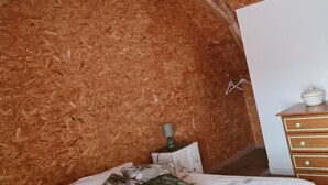 Ein Zimmer mit Holzwänden und einer schrägen Decke, einem Bett mit gemusterten Textilien, einer Kommode mit Schubladen und einem kleinen Tisch mit einer Lampe.