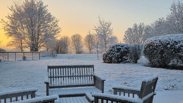 Ein mit Schnee bedeckter Außenbereich mit Sitzgelegenheiten, Bänken und einem Tisch, umgeben von Bäumen und Sträuchern.