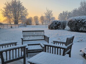 A snow-covered outdoor seating area with benches and a table, surrounded by trees and bushes.