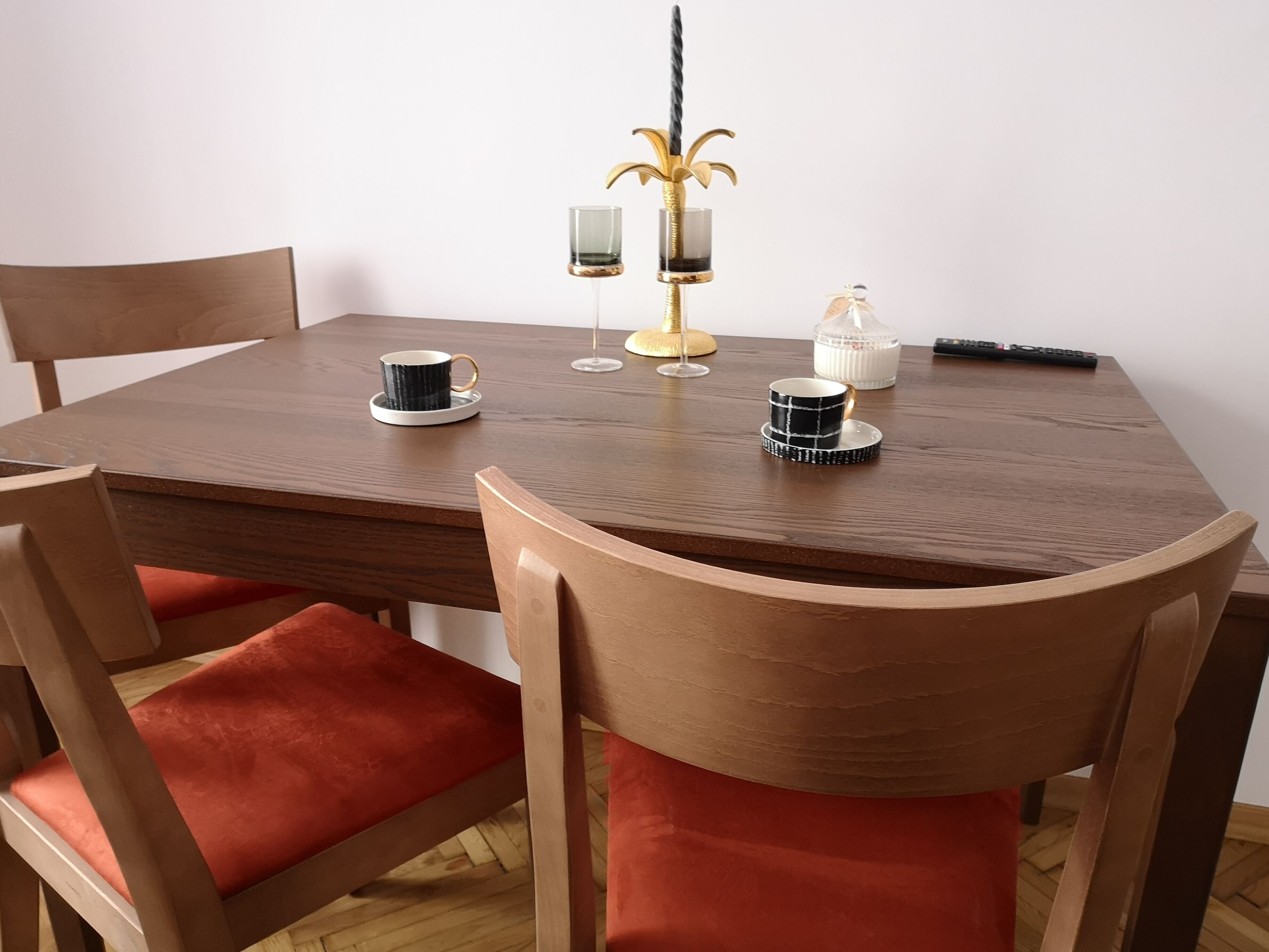 Een houten eettafel met twee stoelen, elk voorzien van een rode kussen. Op de tafel staan twee bekers, een glas, een kandelaar en een afstandsbediening.