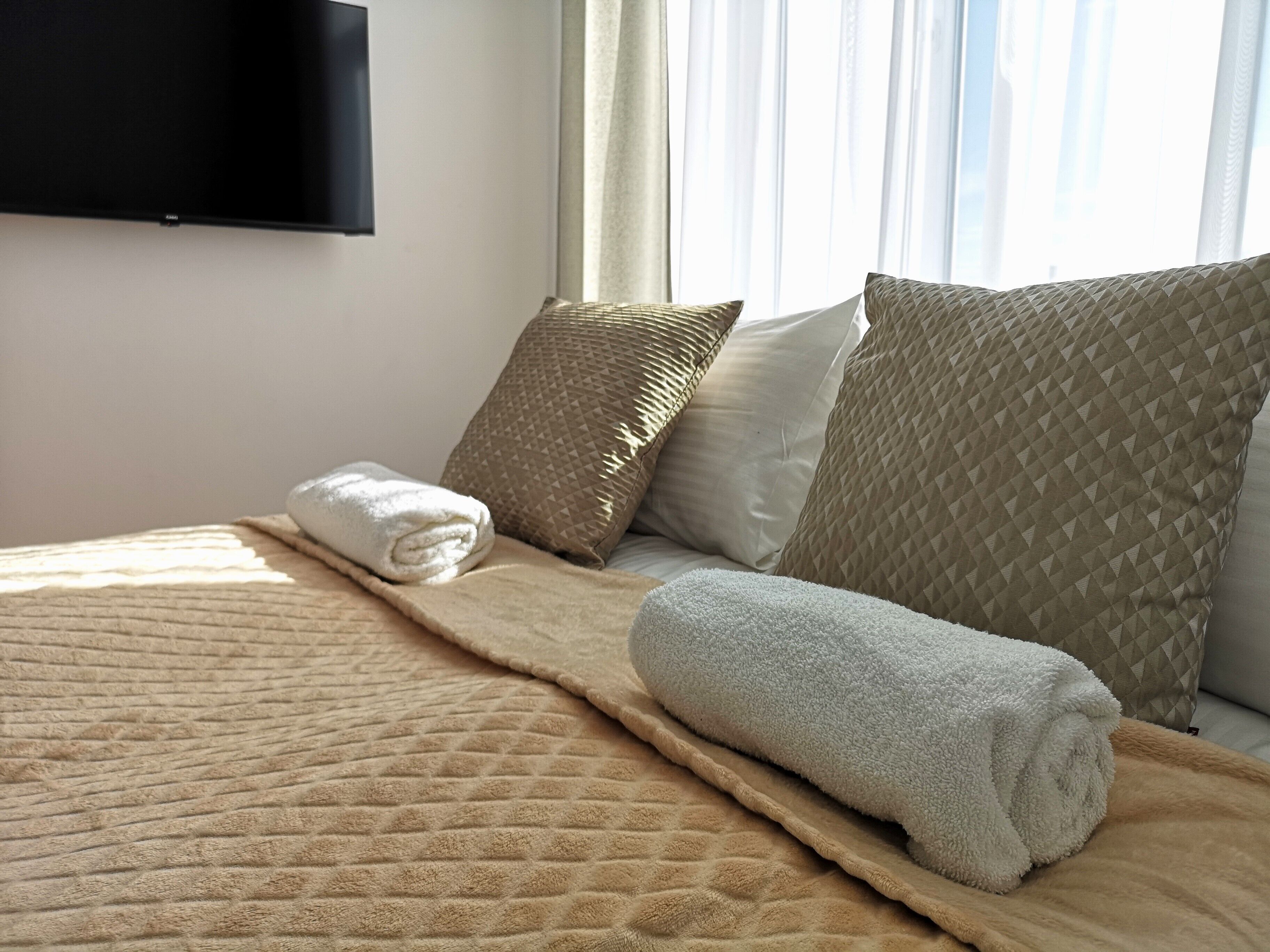 A neatly made bed with a quilted blanket and rolled towels, accompanied by several pillows with geometric patterns.