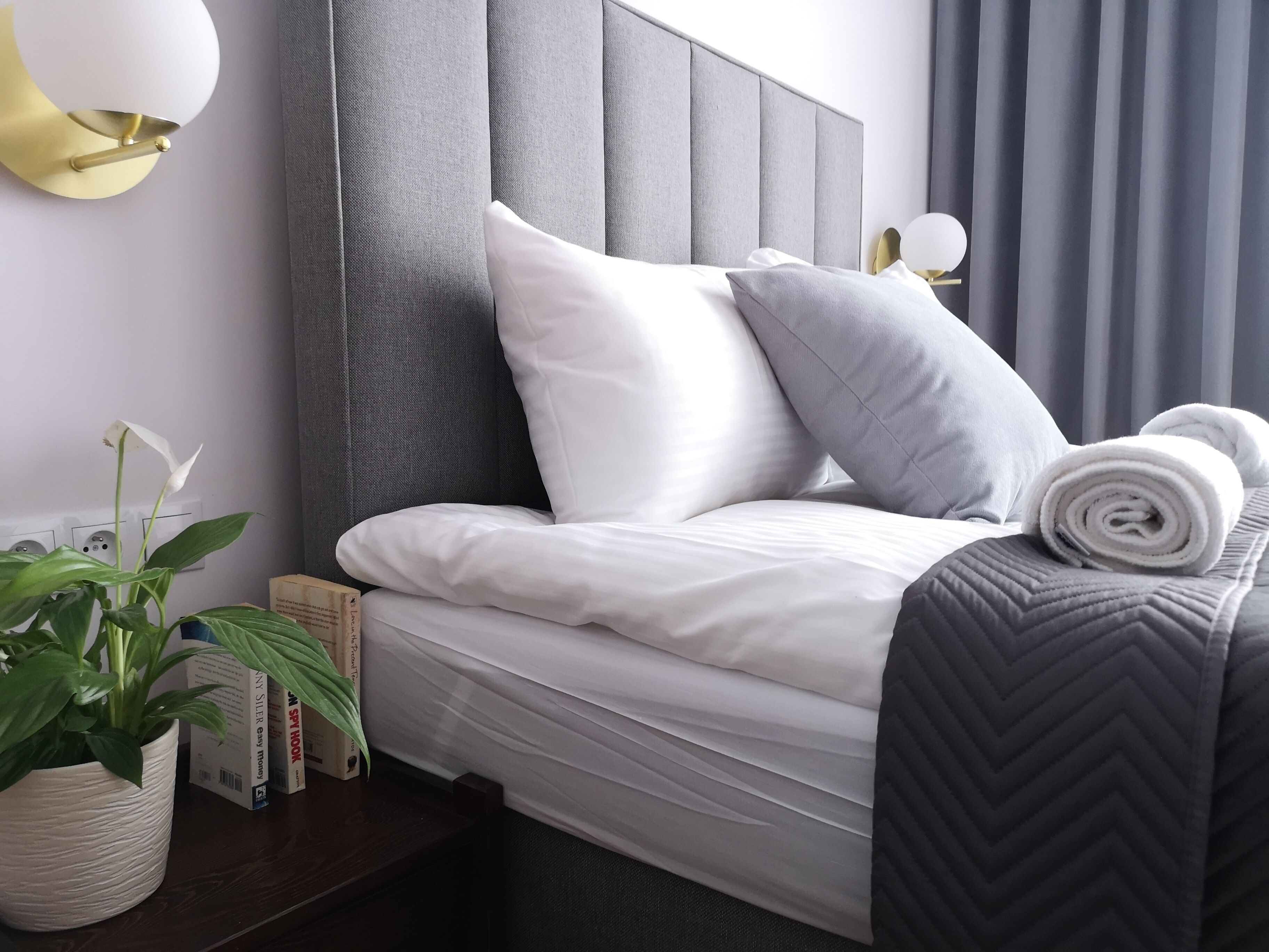 A neatly made bed with white and grey bedding, a grey headboard, and a rolled towel on the side.