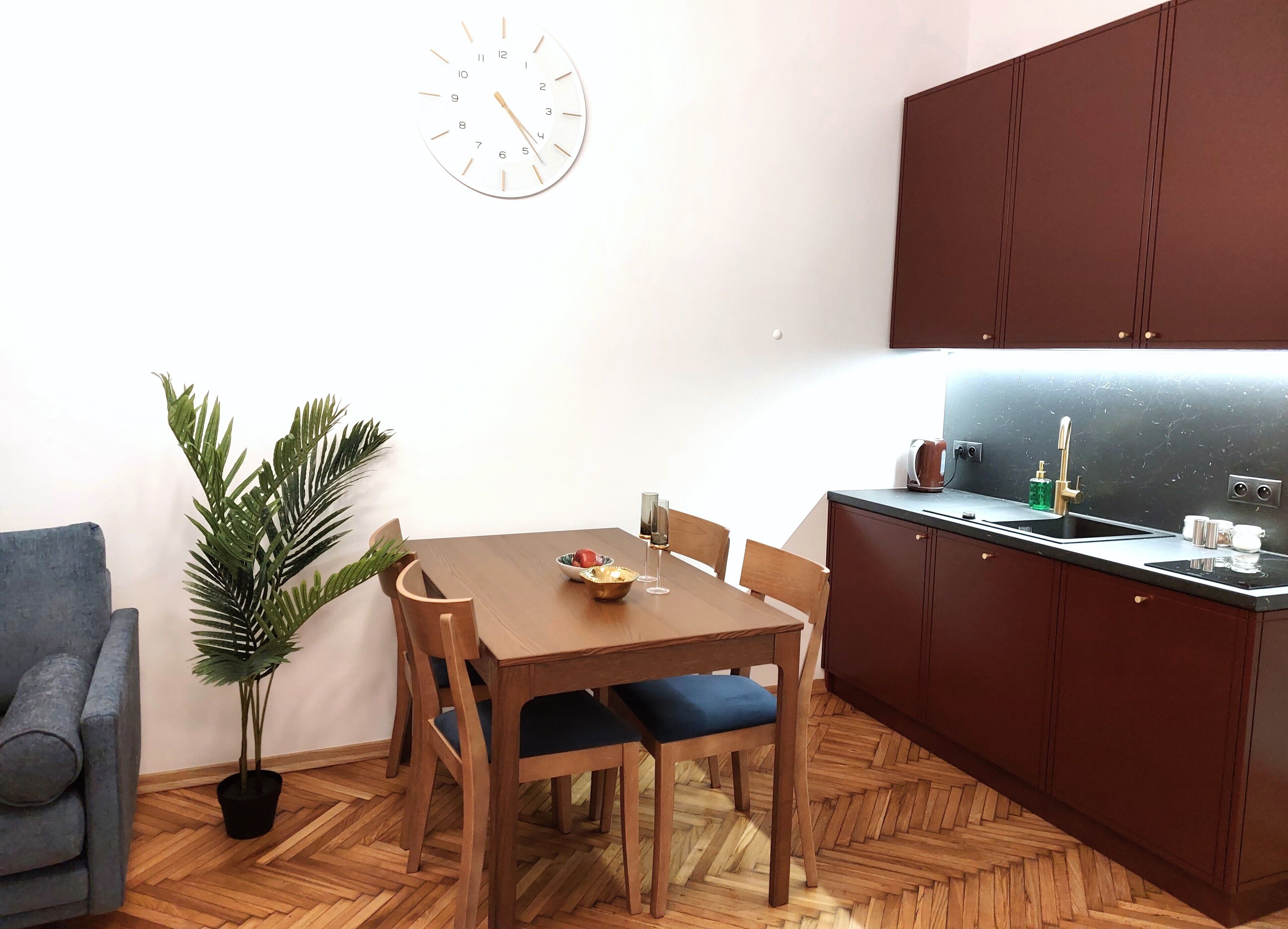 A compact kitchen with a wooden table, chairs, a potted plant, and a clock on the wall.