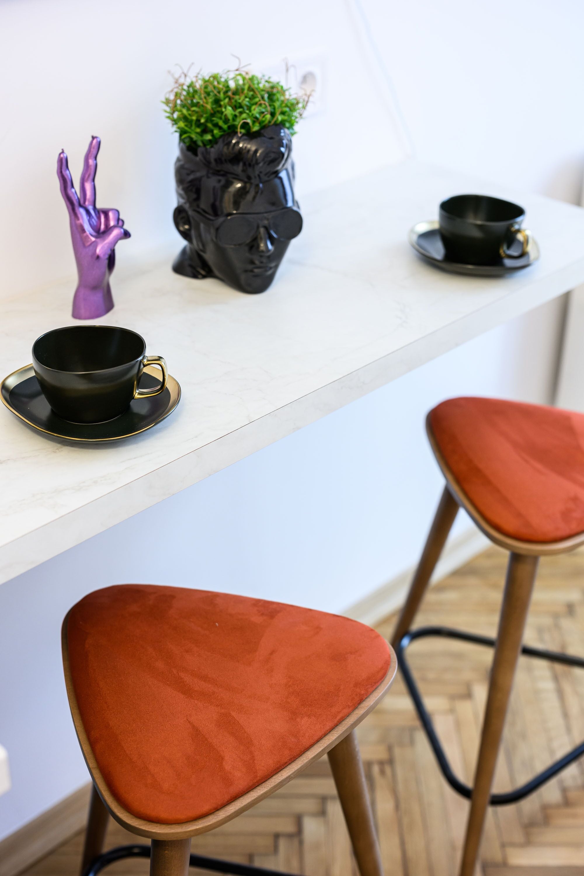 A modern kitchen with a white countertop, two red bar stools, a black coffee cup, a purple hand figurine, and a black pot with a plant.