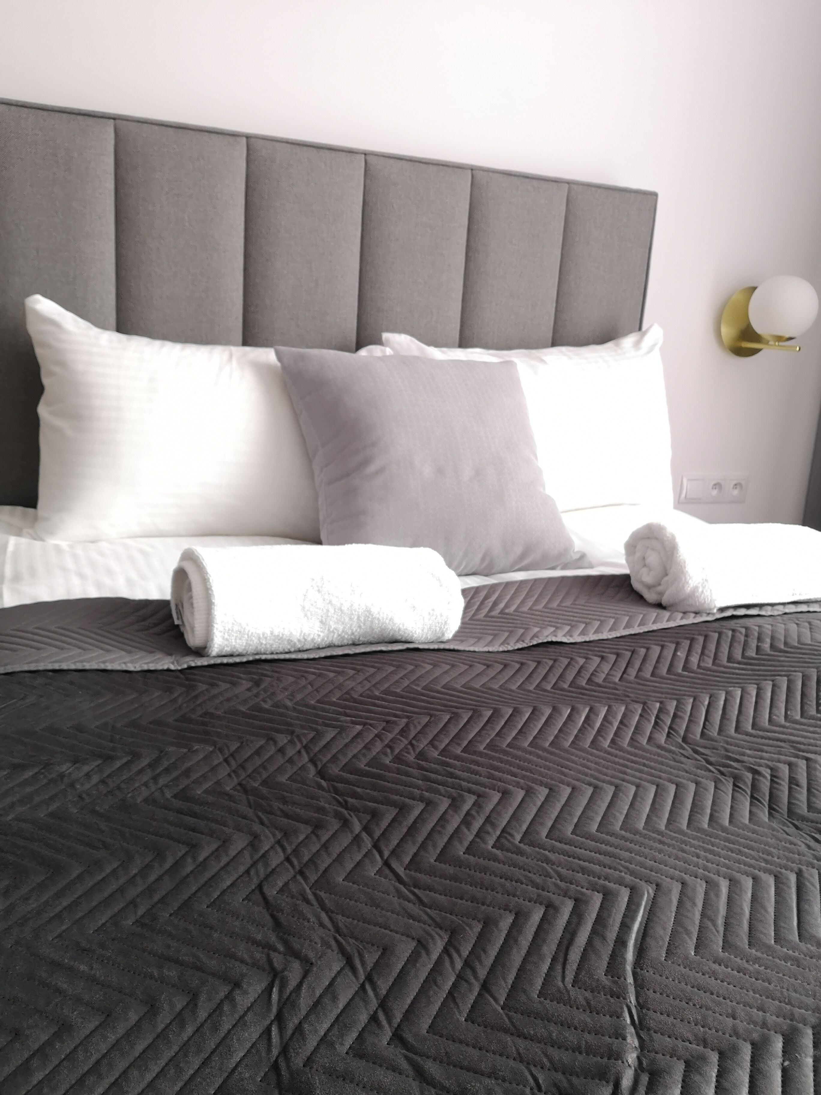 A neatly made bed with a grey quilt, two pillows, and a rolled towel.