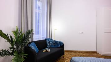 Ein Zimmer mit einer schwarzen Couch, einem blau gesteppten Bett, einer Zimmerpflanze und einem Fenster mit durchsichtigen Vorhängen.