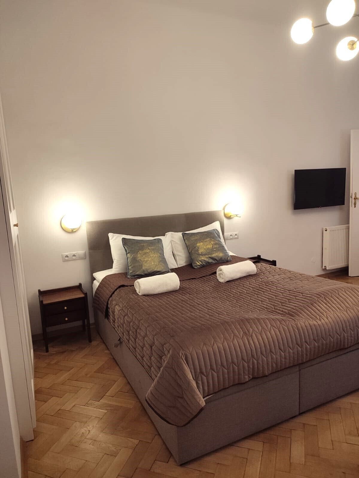 A neatly made bed with a brown quilt, two pillows, and two rolled towels. There is a small bedside table with a lamp, a wall-mounted TV, and a radiator.