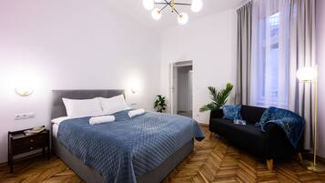 A modern bedroom with a large bed, a black sofa, a wooden herringbone floor, and a ceiling light fixture.