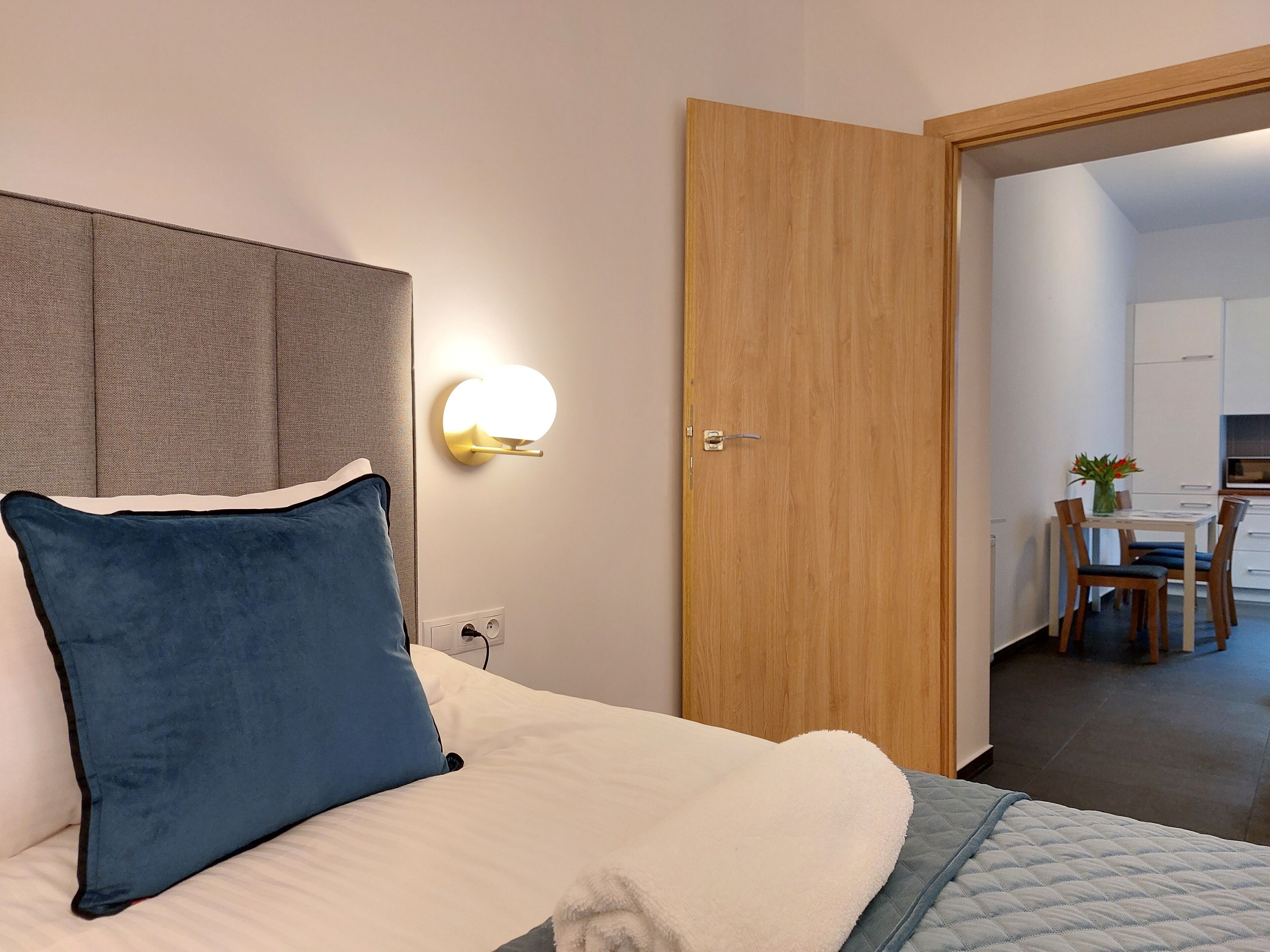 A hotel room with a bed, a wall-mounted light, a wooden door, and a view into the dining area.