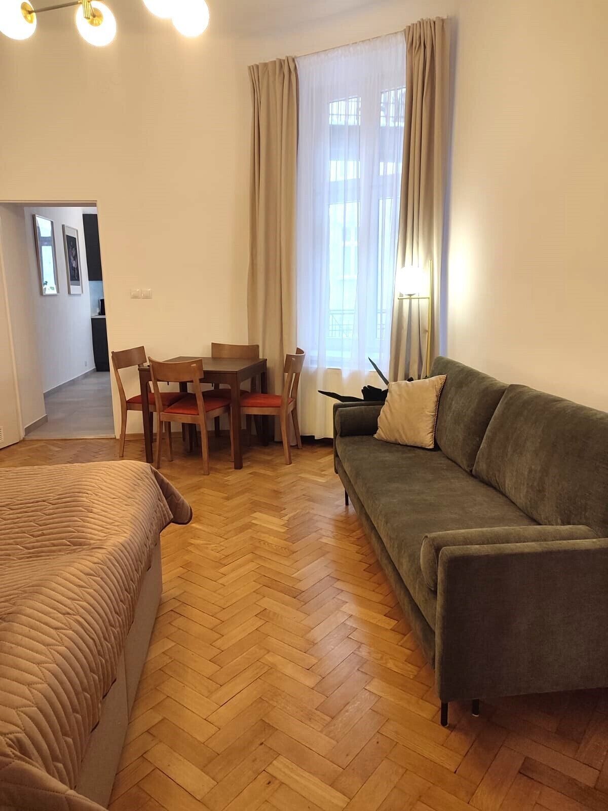 A hotel room with a wooden herringbone floor, a bed, a sofa, a dining table with chairs, and a window with curtains.